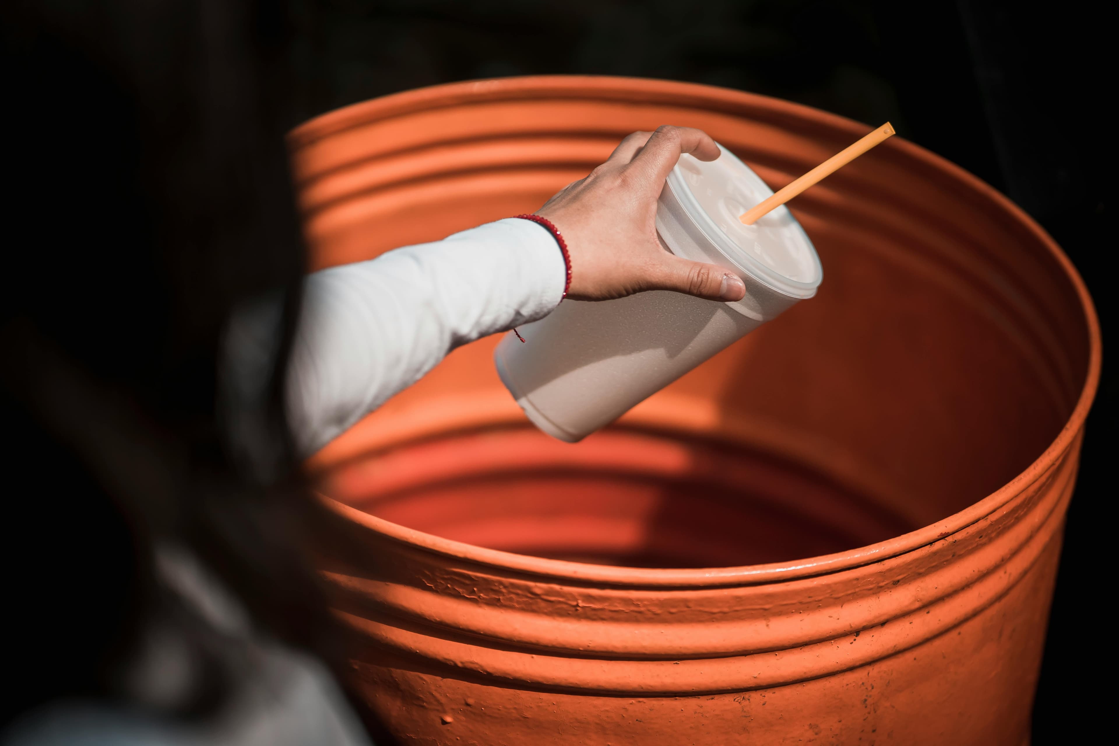 A hand holding a disposable drink cup over an orange bin